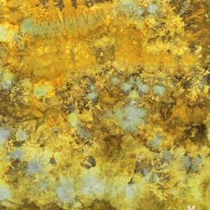 May include: A fabric surface featuring a tie-dye pattern with vibrant shades of golden yellow, mustard, and deep brown, interspersed with small, scattered patches of pale grey and white. The letter M is printed in white in the bottom right corner.