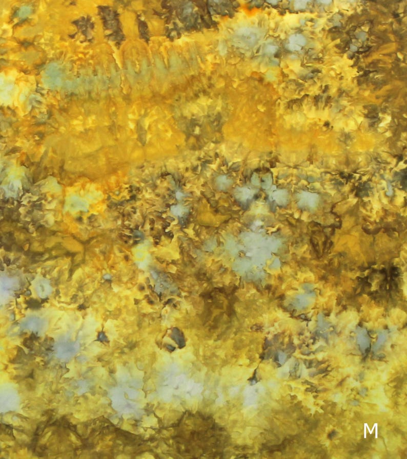May include: A fabric surface featuring a tie-dye pattern with vibrant shades of golden yellow, mustard, and deep brown, interspersed with small, scattered patches of pale grey and white. The letter M is printed in white in the bottom right corner.