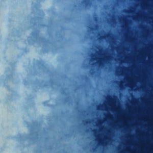 May include: A fabric surface featuring a tie-dye pattern with a gradient transition from light blue on the left to deep navy blue on the right. The design displays organic, cloud-like shapes and dark, branching textures. The letter E is visible.