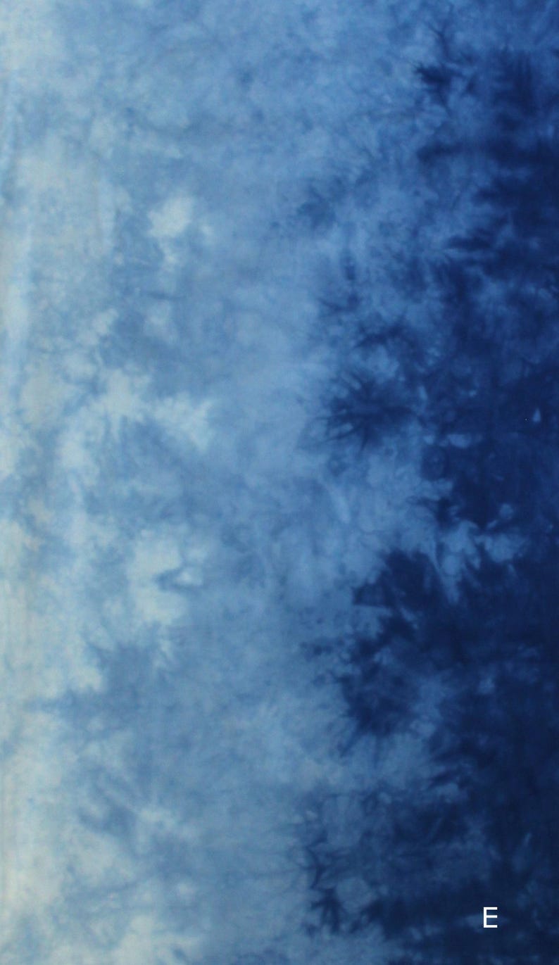 May include: A fabric surface featuring a tie-dye pattern with a gradient transition from light blue on the left to deep navy blue on the right. The design displays organic, cloud-like shapes and dark, branching textures. The letter E is visible.