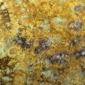 May include: A fabric surface featuring a tie-dye pattern with a blend of golden yellow, mustard, and deep brown hues, interspersed with patches of pale cream and white. The letter P is visible in the bottom right corner of the textile.