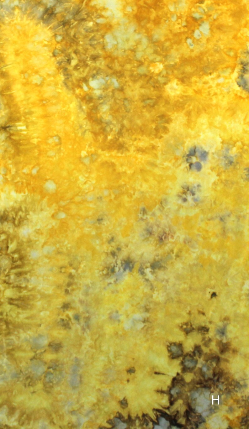 May include: A fabric surface featuring a tie-dye pattern with vibrant shades of yellow, gold, and mustard, accented by dark brown and charcoal gray mottled spots. The letter H is printed in white in the bottom right corner of the textile.