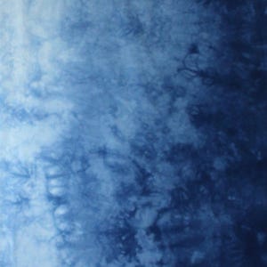 May include: A fabric surface featuring a tie-dye pattern with a gradient transition from light blue on the left to deep navy blue on the right. The texture displays organic, cloud-like shapes and intricate indigo swirls. The letter B is visible.