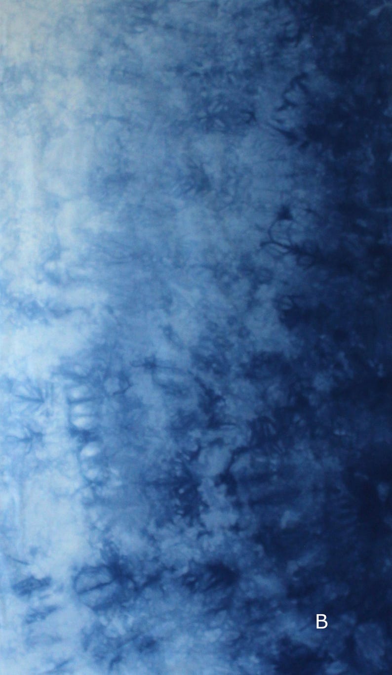 May include: A fabric surface featuring a tie-dye pattern with a gradient transition from light blue on the left to deep navy blue on the right. The texture displays organic, cloud-like shapes and intricate indigo swirls. The letter B is visible.