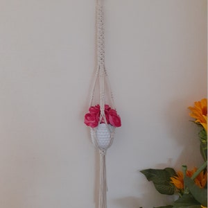 May include: A white macrame plant hanger with a white pot and pink flowers hanging from a hook on a white wall.