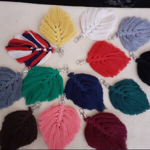 Puede incluir: Una colección de llaveros de plumas de macramé en varios colores, incluyendo amarillo, blanco, rojo, azul, gris, verde, rosa, marrón y negro. Cada llavero cuenta con un cierre plateado para sujetar a llaves o bolsos.