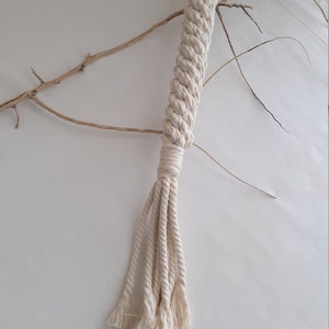 Puede incluir: Un juguete de cuerda de algodón blanco largo, con un mango trenzado y múltiples hebras que cuelgan de la parte inferior. La cuerda está anudada en la parte superior y las hebras están deshilachadas en los extremos.