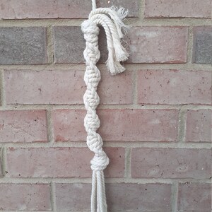 Puede incluir: Un juguete para perros de cuerda de algodón blanco con un diseño anudado. El juguete está colgado de una pared de ladrillo.