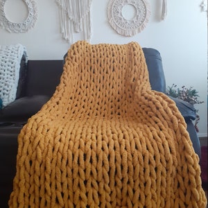 May include: A chunky knit throw blanket in a mustard yellow color. The blanket is draped over a black leather couch.