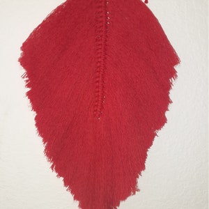 May include: A red macrame wall hanging with a feather-like shape. The hanging is made with a thick, red yarn and has a fringe around the edges.