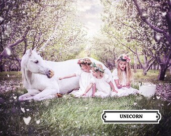 May include: Three girls in white dresses and flower crowns are interacting with a white unicorn in a magical forest setting. The unicorn is lying down in the grass, and the girls are petting it and looking at it. The background is a blur of pink and white flowers.