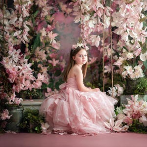 May include: A young person in a pink strapless dress and floral crown sits on a swing surrounded by pink and white flowers. The dress has a layered skirt and a bow in the back. The background is a soft pink and purple.