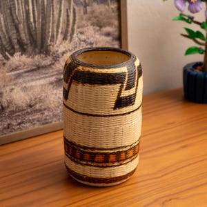 May include: A cylindrical woven basket with a beige base and dark brown and black geometric patterns. The basket has a wide opening and is likely used for storage or decoration. The basket is sitting on a wooden surface.