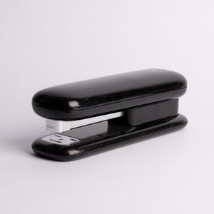 May include: A black stapler with a silver metal component. The stapler has a glossy finish and a rounded design. The image is taken against a white background. The stapler is a common office supply.