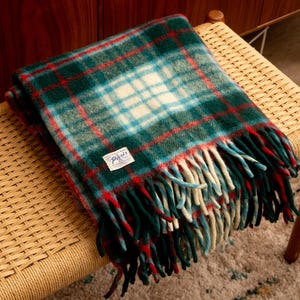 Puede incluir: Una manta plegada a cuadros verdes, rojos y blancos con bordes con flecos. La manta tiene una etiqueta que dice "Paris". La manta está sobre un banco tejido.