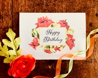 Könnte beinhalten: Eine weiße rechteckige Grußkarte mit einem Aquarell-Blumenkranz in Rot- und Grüntönen, der den Text Happy Birthday in schwarzer Schreibschrift umrahmt. Eine einzelne rote Kunstblume und ein dünnes orangefarbenes und gelbes Band sind zu sehen.