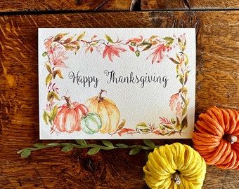 May include: A rectangular white greeting card featuring the text Happy Thanksgiving in black script. The card is decorated with a watercolor border of autumn leaves and three pumpkins in shades of orange, yellow, and green.