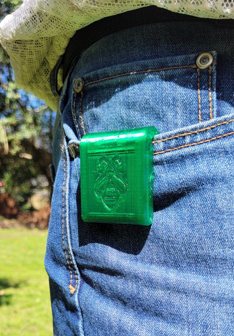 Puede incluir: Un accesorio rectangular verde brillante con un emblema de abeja est&aacute; sujeto a un bolsillo de mezclilla azul. El dise&ntilde;o de la abeja est&aacute; grabado en la superficie. El accesorio est&aacute; hecho de un material s&oacute;lido, posiblemente pl&aacute;stico.
