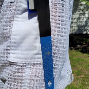 Puede incluir: Una herramienta para colmenas negra y azul con una cuchilla plateada est&aacute; metida en un bolsillo blanco. La herramienta tiene un mango azul con un patr&oacute;n de panal y el texto "THE HONEYSTEAD" con un gr&aacute;fico de abeja.