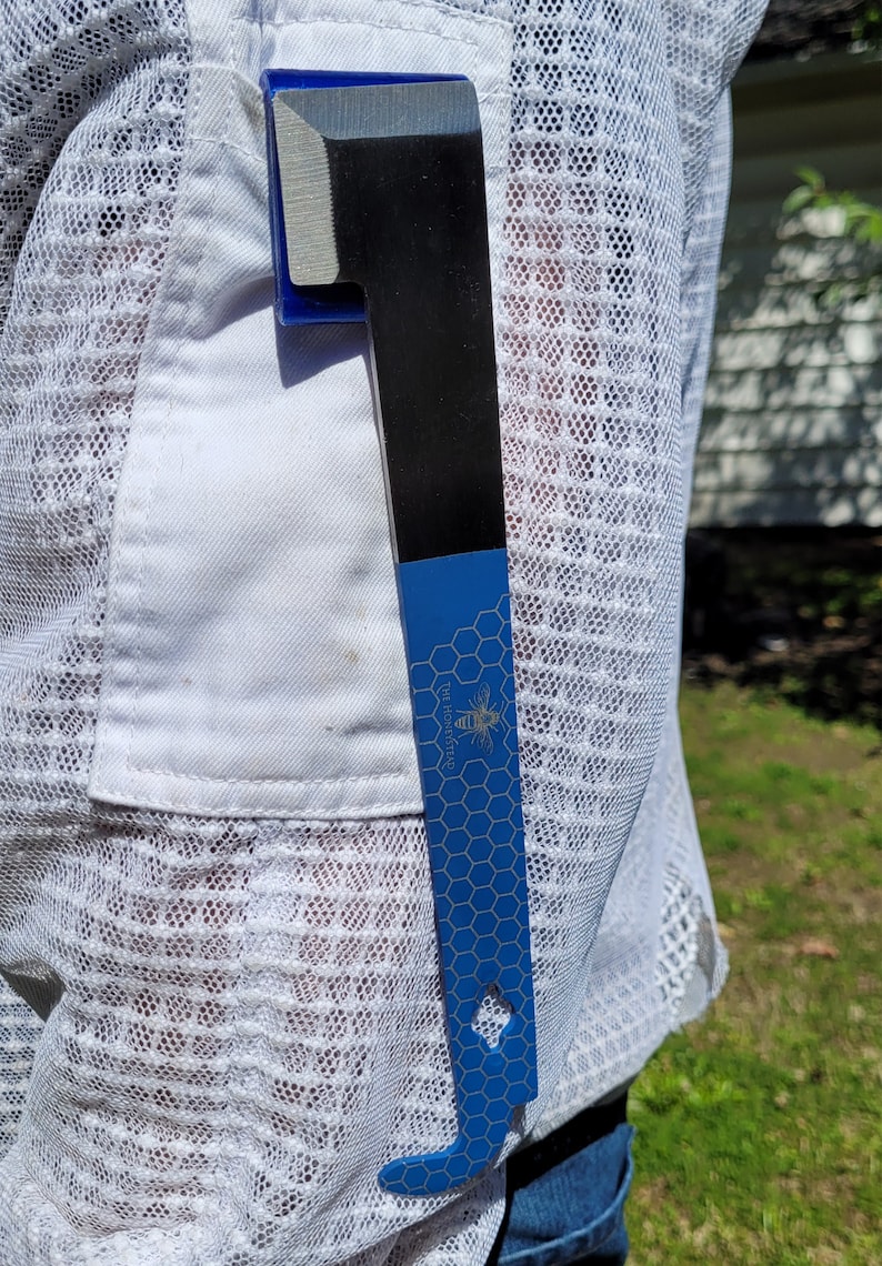 Puede incluir: Una herramienta para colmenas negra y azul con una cuchilla plateada est&aacute; metida en un bolsillo blanco. La herramienta tiene un mango azul con un patr&oacute;n de panal y el texto "THE HONEYSTEAD" con un gr&aacute;fico de abeja.