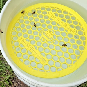 Puede incluir: Un bebedero de abejas de plástico amarillo con diseño de panal flota en un cubo blanco con agua. Varias abejas son visibles en el bebedero. El texto "The Bee Barge" y "The3DBee.com" están impresos en el bebedero.