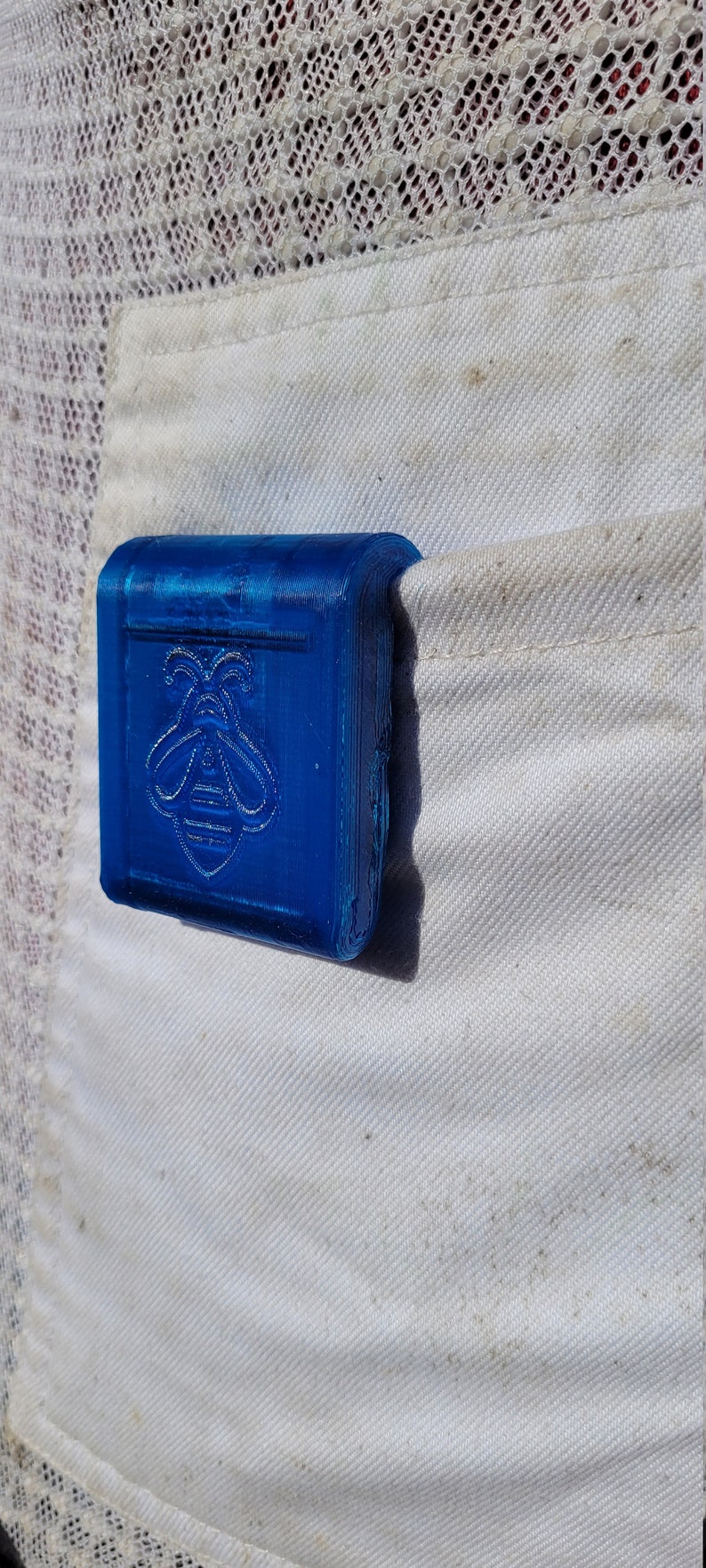 Puede incluir: Un objeto rectangular azul brillante con un dise&ntilde;o de abeja se coloca en un bolsillo blanco. El dise&ntilde;o de la abeja est&aacute; en relieve en la parte delantera. Probablemente sea un peque&ntilde;o contenedor o accesorio.