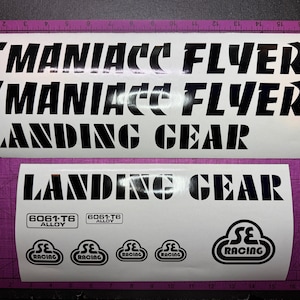 May include: Two white sticker sheets with black text. The top sheet reads "MANIACC FLYER" twice and "LANDING GEAR." The bottom sheet reads "LANDING GEAR," "6061-T6 ALLOY," and "SE RACING" logos. The stickers are on a purple surface.