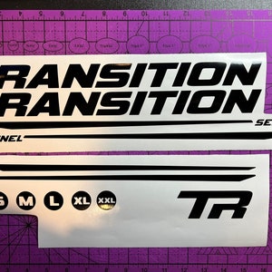 May include: Black and white sticker set for a bicycle. The stickers feature the words "Transition" and "Sentinel" along with size options "S, M, L, XL, XXL" and the letters "TR".