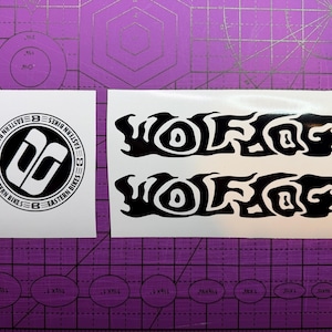 May include: Two white vinyl stickers on a purple cutting mat. One sticker features a black and white circular logo with the text "EASTERN BIKES". The other sticker has the word "WOLFOG" in black, repeated twice.