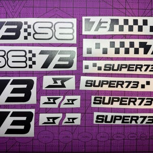 Puede incluir: Una colección de calcomanías en blanco y negro con los números "73" y el texto "SUPER73" en varias fuentes y diseños. Algunas calcomanías incluyen patrones de cuadros y logotipos estilizados. Las calcomanías están dispuestas sobre una esterilla de corte morada.