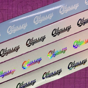 May include: Close-up of four rolls of sticker tape with the word "Odyssey" printed in different colors and styles. The stickers are white, black, iridescent, and holographic.
