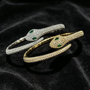 Può includere: Due bracciali rigidi a forma di serpente, uno in metallo color argento e uno in color oro. Entrambi i bracciali sono tempestati di piccoli cristalli trasparenti e presentano due piccole pietre verdi taglio marquise come occhi.