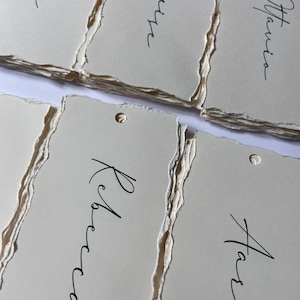 May include: A stack of white, textured paper tags with handwritten names on each tag: Octavia, Louise, Beth, Rebecca, and Harrison.