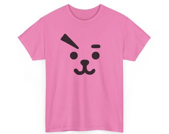 May include: A short-sleeved pink t-shirt featuring a black graphic print of a stylized animal face. The design includes two round eyes, a small nose, a curved mouth, and a single slanted eyebrow above the right eye, centered on the chest.