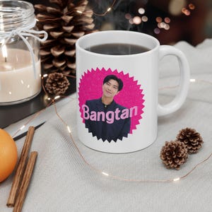 Può includere: Tazza in ceramica bianca con un design a stella rosa, una foto di una persona e la parola "Bangtan" in rosa. La tazza contiene un liquido scuro e ha un manico curvo. La tazza è su una superficie grigio chiaro.