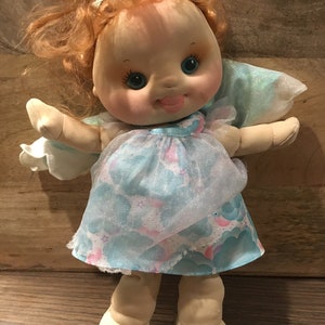 May include: A vintage doll with red hair, blue eyes, and a pink tongue sticking out. She is wearing a light blue dress with a white and pink cloud pattern and a sheer blue overlay. The doll has a white headband and is standing on a wooden surface.