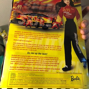 May include: Barbie doll dressed in a red and yellow NASCAR racing uniform with the number 94 on the front. The doll is holding a McDonald's drive-thru cup. The background is a checkered black and white pattern.