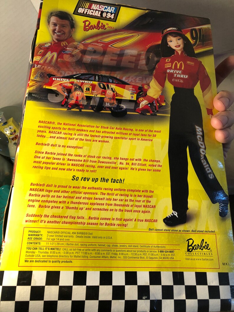May include: Barbie doll dressed in a red and yellow NASCAR racing uniform with the number 94 on the front. The doll is holding a McDonald's drive-thru cup. The background is a checkered black and white pattern.
