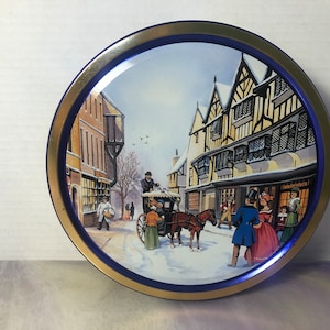 May include: A round tin with a blue and gold rim. The tin features a winter scene with a horse-drawn carriage, people walking down a snowy street, and buildings with snow-covered roofs.