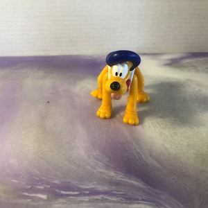 Op de afbeelding: Een gele plastic speelgoedhond met een blauwe hoed. De hond heeft een rode neus en zwarte ogen. Het speelgoed zit op een paarse en witte ondergrond.