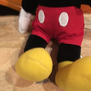 Może przedstawiać: Pluszowa zabawka Mickey Mouse z czarnym futrem, czerwonymi spodenkami z białymi k&oacute;łkami i ż&oacute;łtymi butami.