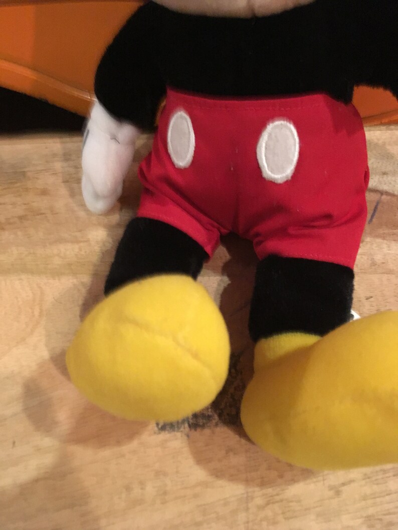 Może przedstawiać: Pluszowa zabawka Mickey Mouse z czarnym futrem, czerwonymi spodenkami z białymi k&oacute;łkami i ż&oacute;łtymi butami.