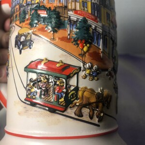 K&ouml;nnte beinhalten: Eine wei&szlig;e Keramiktasse mit rotem Rand und einer bunten Illustration einer Stra&szlig;enszene in einem Themenpark. Die Illustration zeigt einen roten und gr&uuml;nen Trolley mit Mickey Mouse und anderen Disney-Charakteren, die darin fahren. Eine Pferdekutsche zieht den Trolley. Der Text "Refreshment Corner" ist auf einem Geb&auml;ude im Hintergrund zu sehen. Die Tasse tr&auml;gt den Text "Disney World" am Boden.