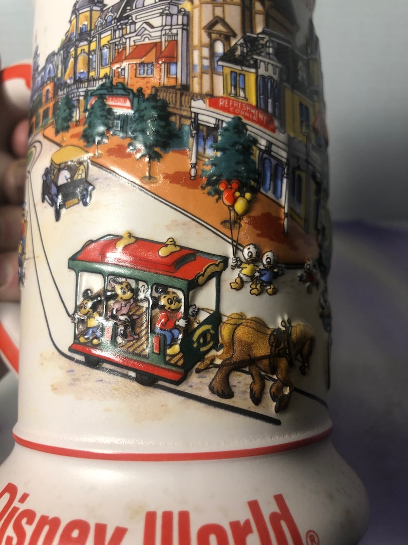 K&ouml;nnte beinhalten: Eine wei&szlig;e Keramiktasse mit rotem Rand und einer bunten Illustration einer Stra&szlig;enszene in einem Themenpark. Die Illustration zeigt einen roten und gr&uuml;nen Trolley mit Mickey Mouse und anderen Disney-Charakteren, die darin fahren. Eine Pferdekutsche zieht den Trolley. Der Text "Refreshment Corner" ist auf einem Geb&auml;ude im Hintergrund zu sehen. Die Tasse tr&auml;gt den Text "Disney World" am Boden.