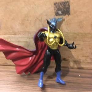 May include: A blue and gold action figure with a red cape. The figure has a blue face and a gold chest piece.
