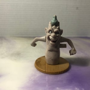 May include: A gray plastic toy figure of a troll with a small bird on its head. The troll has a wide smile and is standing on a brown base.