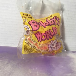 May include: A yellow and pink plastic bag with the words "Happy Meal" and "Bobby's World" printed on it. The bag contains a pink and white plastic toy that looks like a spaceship.