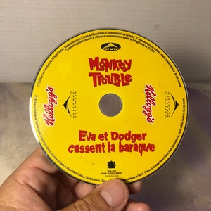 Może przedstawiać: Żółta płyta DVD z tytułem "Monkey Trouble" i tekstem "Eva et Dodger cassent la baraque" w kolorze czerwonym. Płyta jest oznaczona logo Kellogg's i logo New Line Home Entertainment.