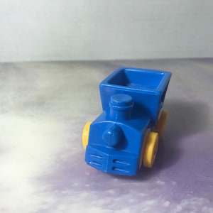 May include: A blue toy train with yellow wheels. The train has a boxcar on the back.