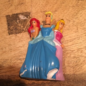 May include: Three plastic Disney princess figurines. The princess in the center is Cinderella, wearing a blue dress and a tiara. The princess on the left is Ariel, wearing a red top and a green tail. The princess on the right is Belle, wearing a pink dress and a yellow crown.